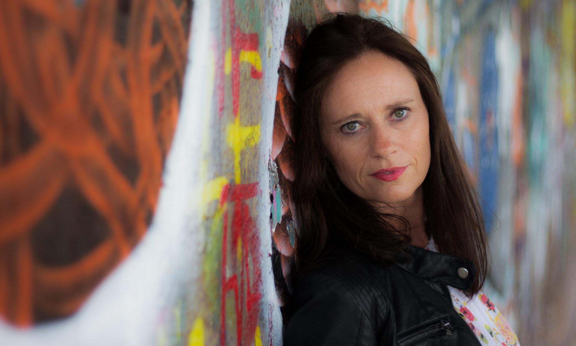 Portrait einer dunkelhaarigen Frau in schwarzer Jacke vor einer bunten Graffitiwand. Sie blickt direkt in die Kamera.