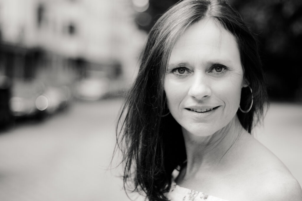 Schwarz-Weiß-Portrait einer Frau mit dunklen, schulterlangen Haaren und Ohrringen. Sie blickt direkt in die Kamera, während der städtische Hintergrund unscharf bleibt.