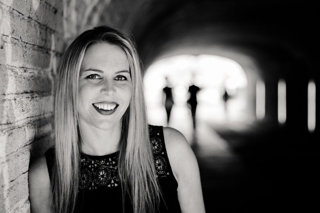 Schwarz-Weiß-Portrait einer lächelnden Frau mit langen blonden Haaren und schwarzem Oberteil. Sie lehnt an einer Backsteinwand. Im unscharfen Hintergrund sind die Silhouetten zweier Personen in einem Tunnel zu sehen.