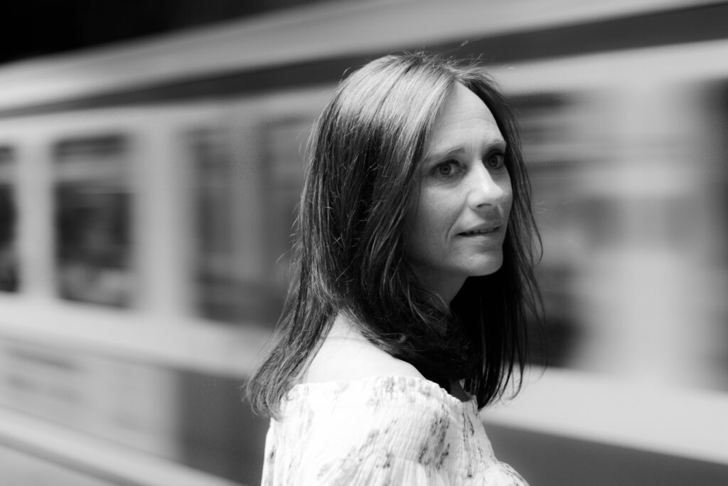 Schwarz-Weiß-Portrait einer dunkelhaarigen Frau in heller Kleidung. Sie blickt seitlich über ihre Schulter, während im unscharfen Hintergrund eine U-Bahn zu sehen ist.