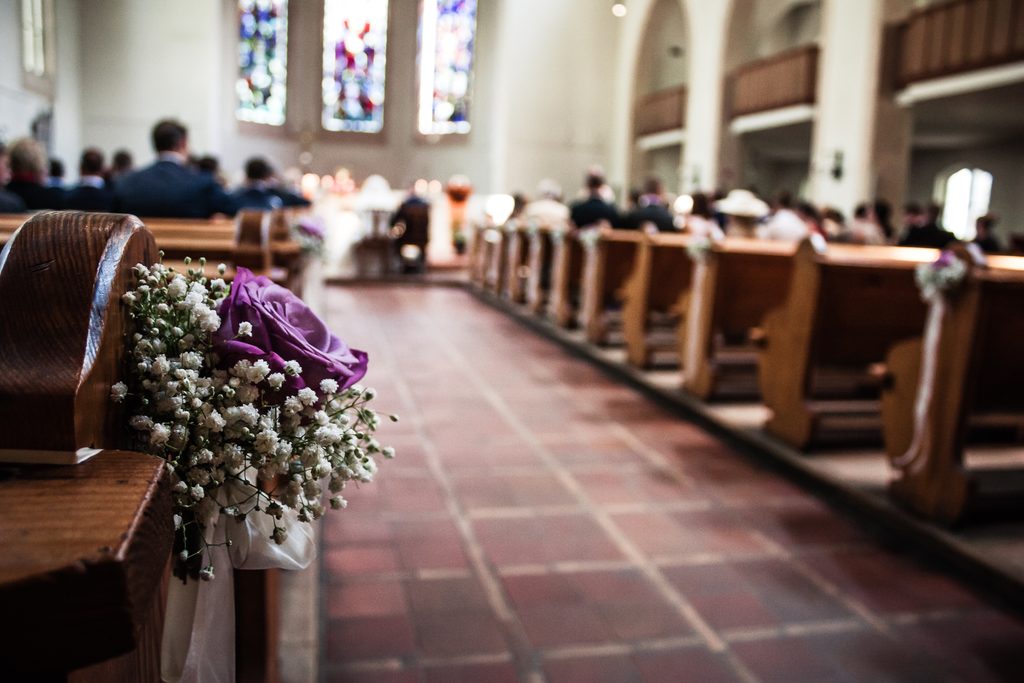 Blumenschmuck mit einer violetten Rose und weißem Schleierkraut an einer Kirchenbank. Im unscharfen Hintergrund sitzen Hochzeitsgäste während einer Trauung in der Kirche.