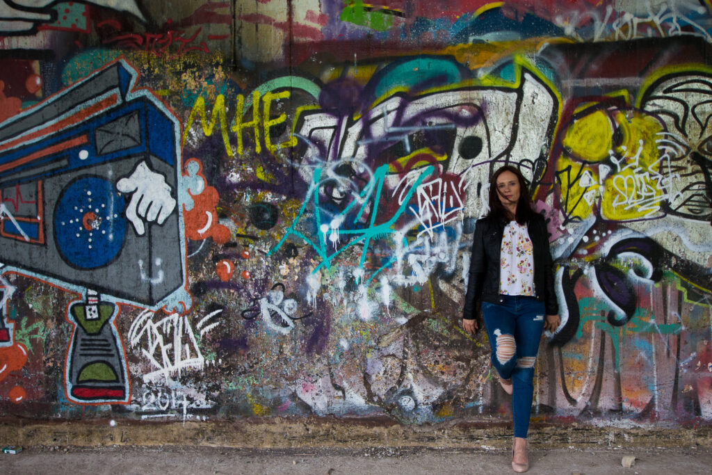 Eine Frau mit dunklen Haaren, schwarzer Jacke und blauer Jeans steht vor einer großflächigen, bunten Graffitiwand mit unterschiedlichen Schriftzügen und Motiven.